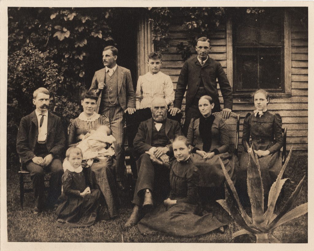 The Morley family. An old family portrait; ten people stand in front of a clapboard house. Lucy Martindale and Thomas Morley are in the center. 
